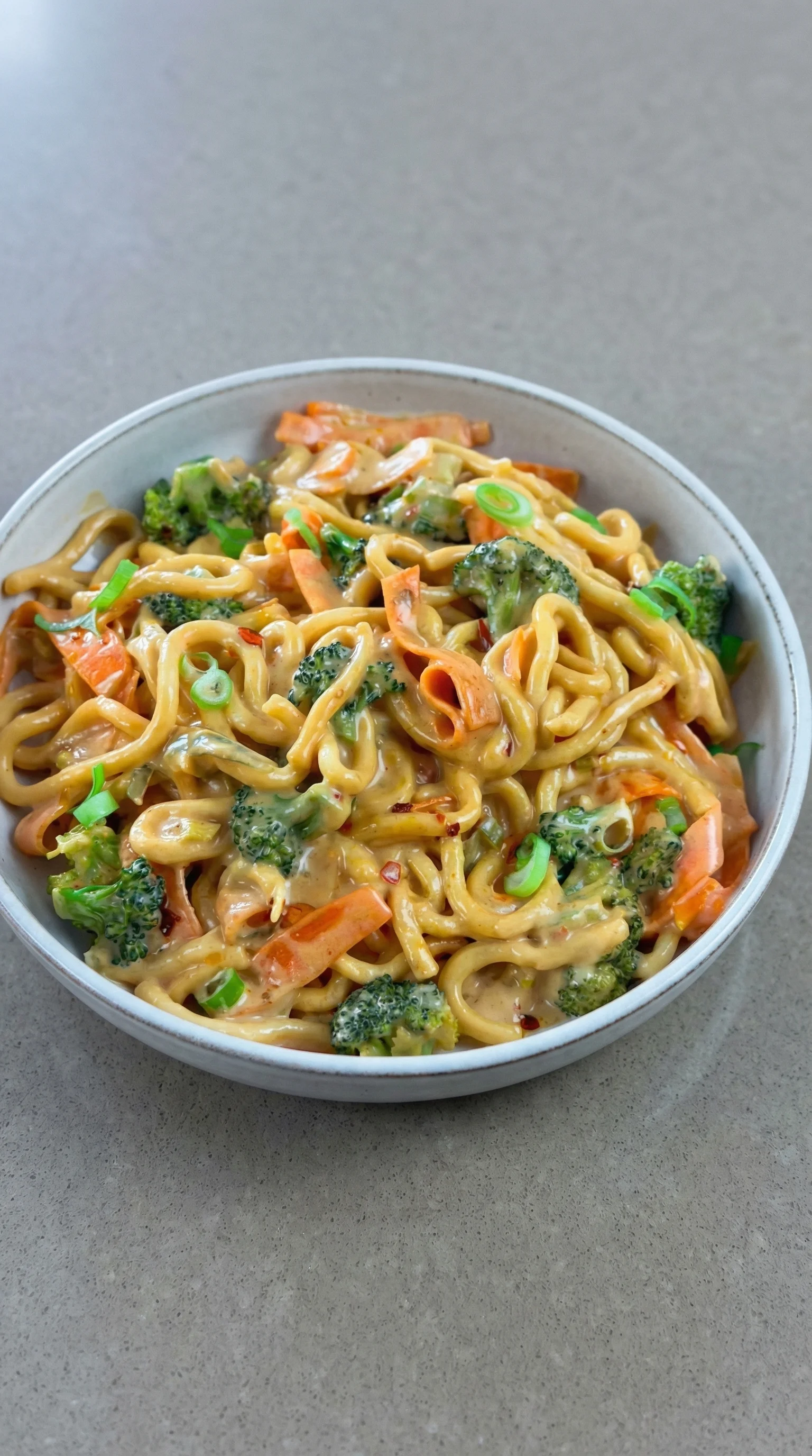Ein weißer Teller mit Udon Nudeln, die mit Erdnusssauce und Gemüse garniert sind.